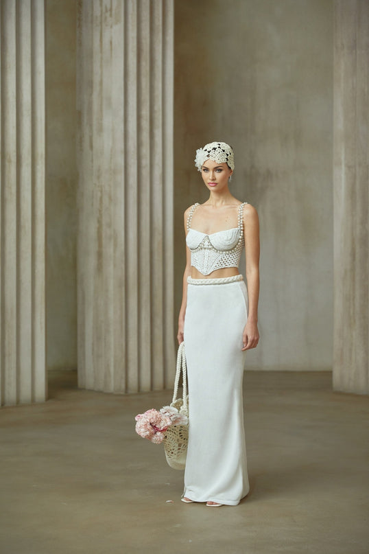 A woman wearing a white crochet knit two piece outfit consisting of a sleeveless crop top and matching high waisted mini skirt with a scalloped hem