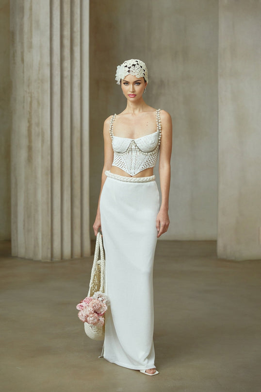 A woman wearing a white crochet knit two piece set consisting of a cropped top and midi skirt with intricate floral patterns standing against a plain background