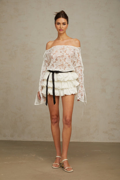 A woman wearing a white floral lace matching set consisting of a cropped top and skirt standing against a plain background