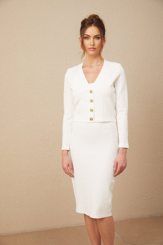 A woman wearing a white knit dress with a monogram pattern standing against a plain background