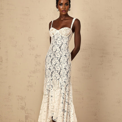 A woman wearing a white maxi dress with floral lace detailing and a bustier bodice standing against a plain background