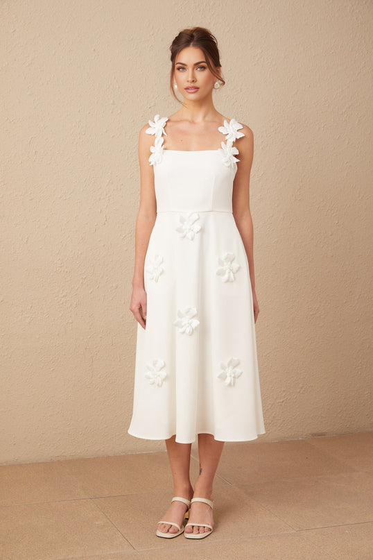 A woman wearing a white midi dress with a square neckline adorned with faux flower details standing against a neutral background