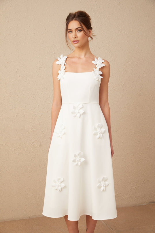A woman wearing a white midi dress with a square neckline adorned with faux flower details standing against a plain background