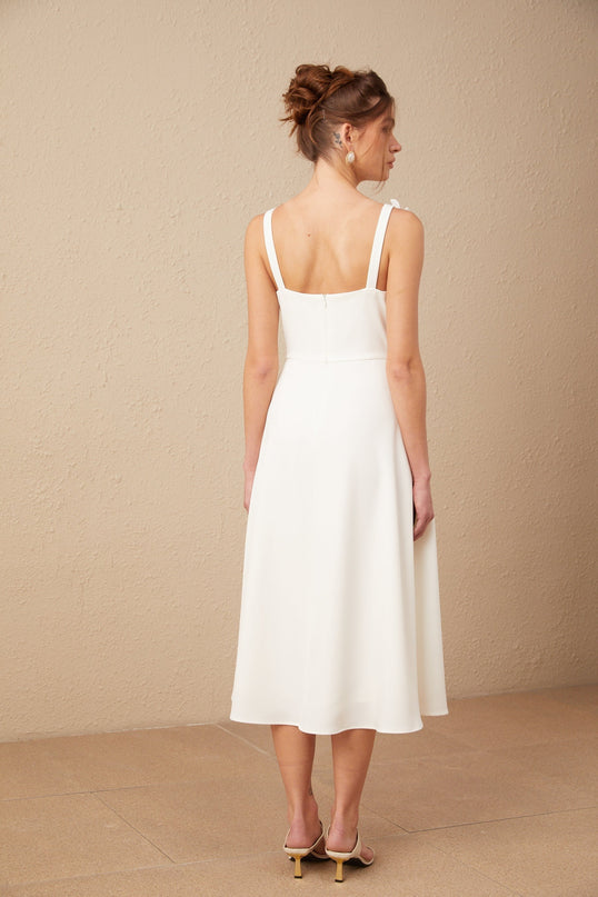 A woman wearing a white midi dress with a square neckline adorned with faux flower details standing against a plain background