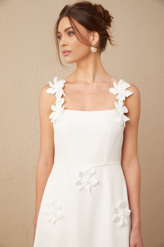 A woman wearing a white midi dress with a square neckline and faux flower details standing against a plain background