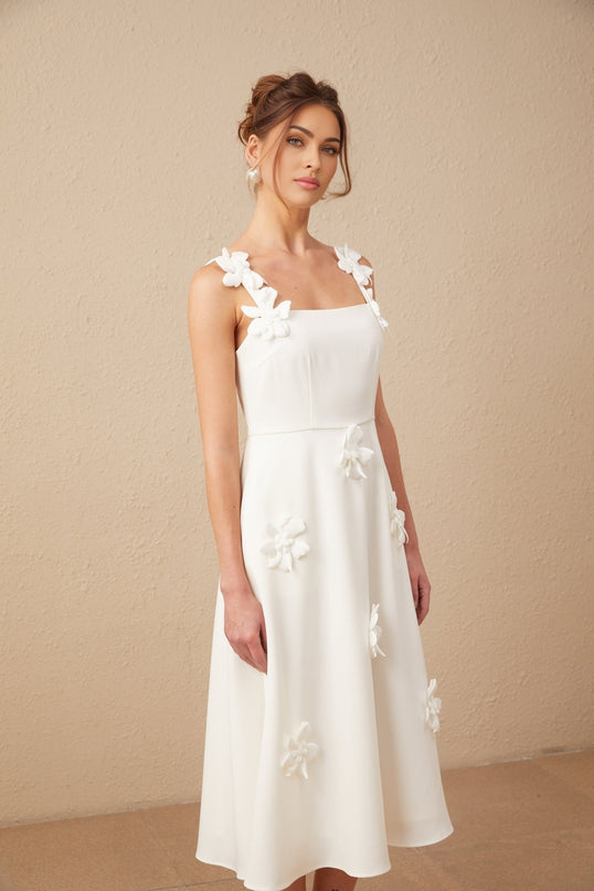 A woman wearing a white midi dress with a square neckline and faux flower details standing against a plain background