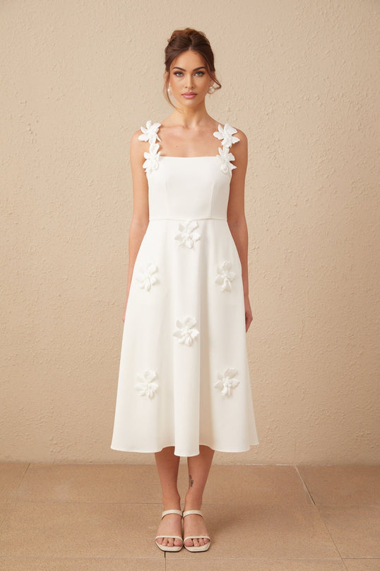 A woman wearing a white midi dress with a square neckline and faux flower embellishments standing in a relaxed pose