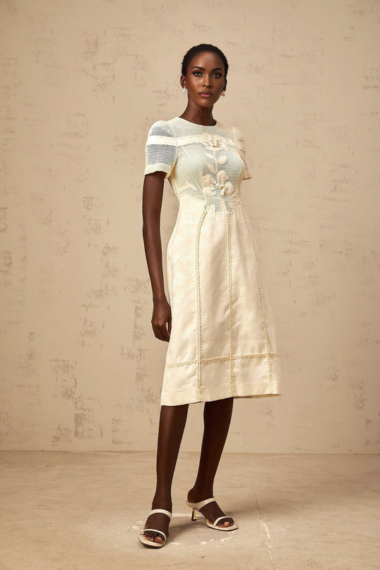 A woman wearing a white midi dress with colorful floral embroidery and openwork detailing standing in a neutral background