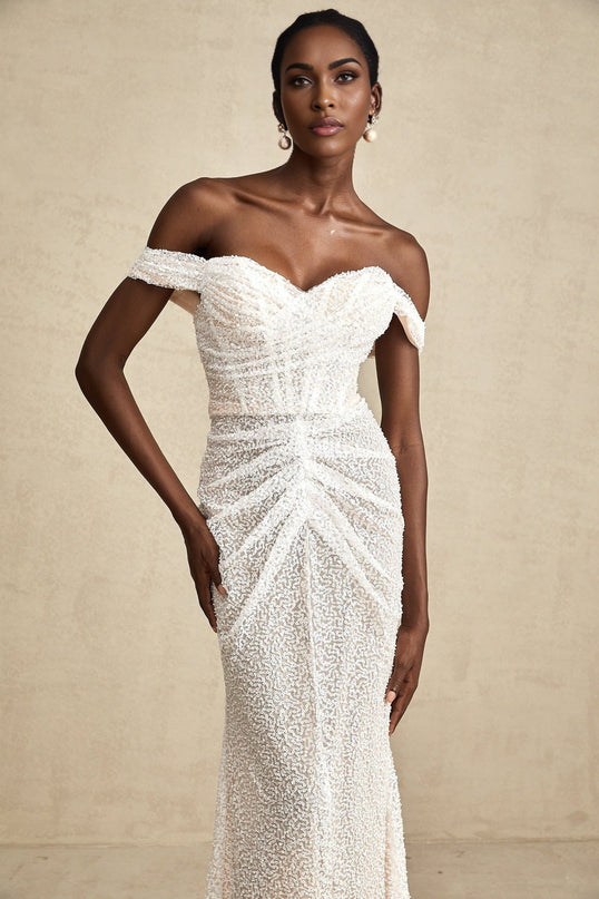 A woman wearing a white offshoulder maxi dress with embellishments standing against a plain background