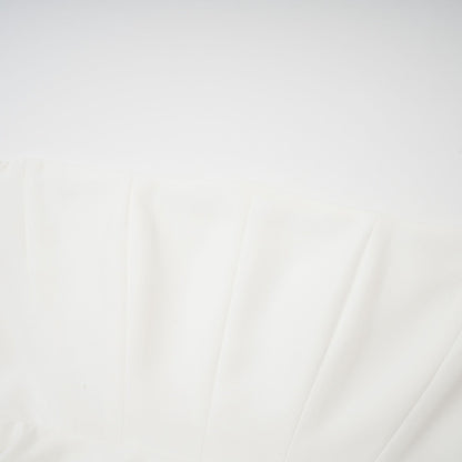 A woman wearing a white offshoulder maxi dress with pleated details standing against a plain background
