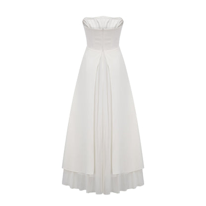 A woman wearing a white pleated offshoulder maxi dress with a flowing skirt standing against a plain background