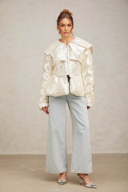 A woman wearing a white puffy down coat covered in sparkling sequins standing against a plain background