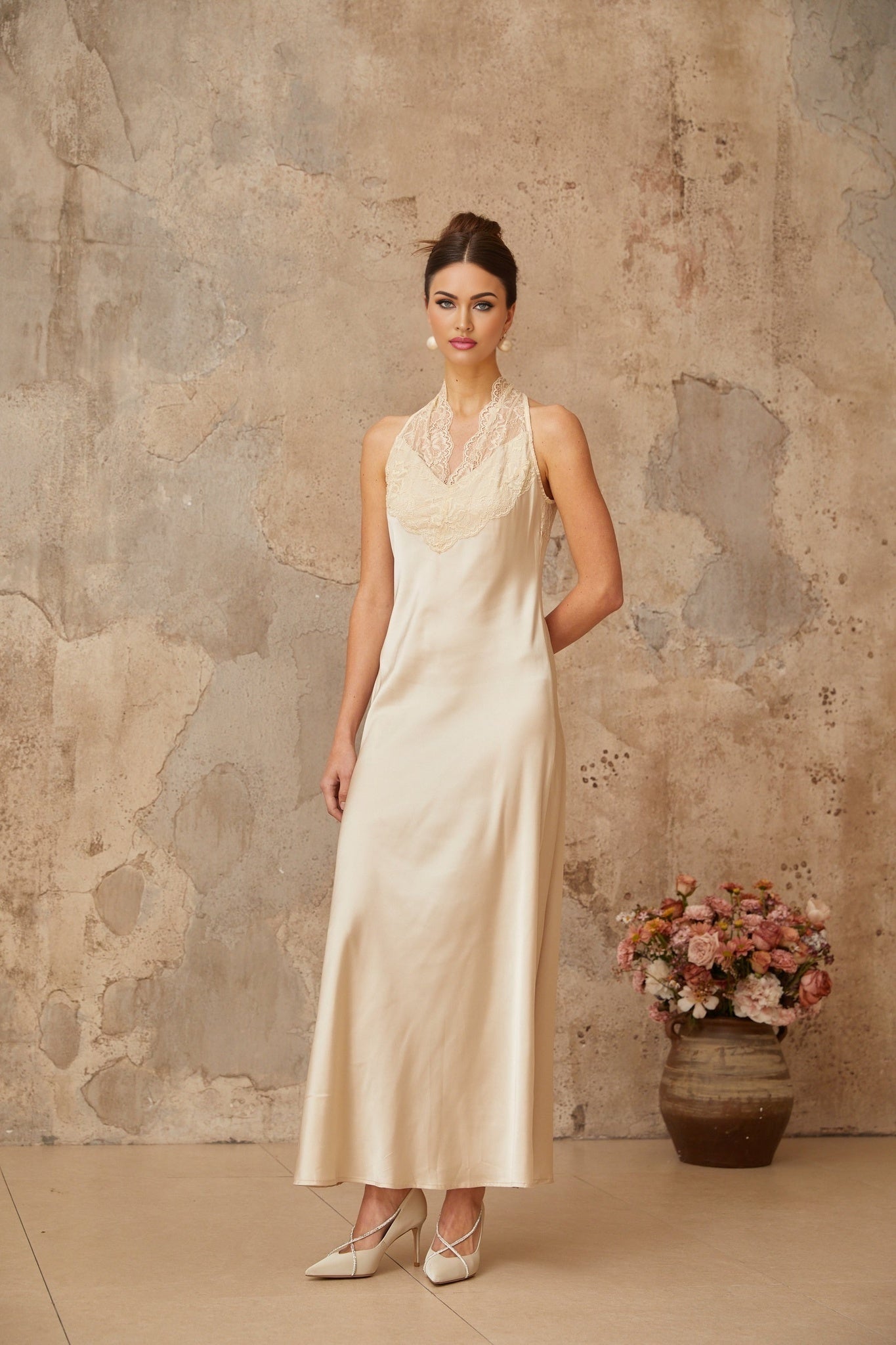 A woman wearing a white satin maxi dress with lace trim standing against a plain background