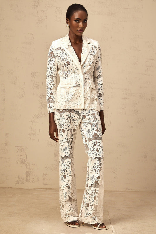 A woman wearing a white tulle blazer and matching trousers adorned with sequined flower embroidery The set features delicate floral patterns shimmering with sequins against a sheer tulle background