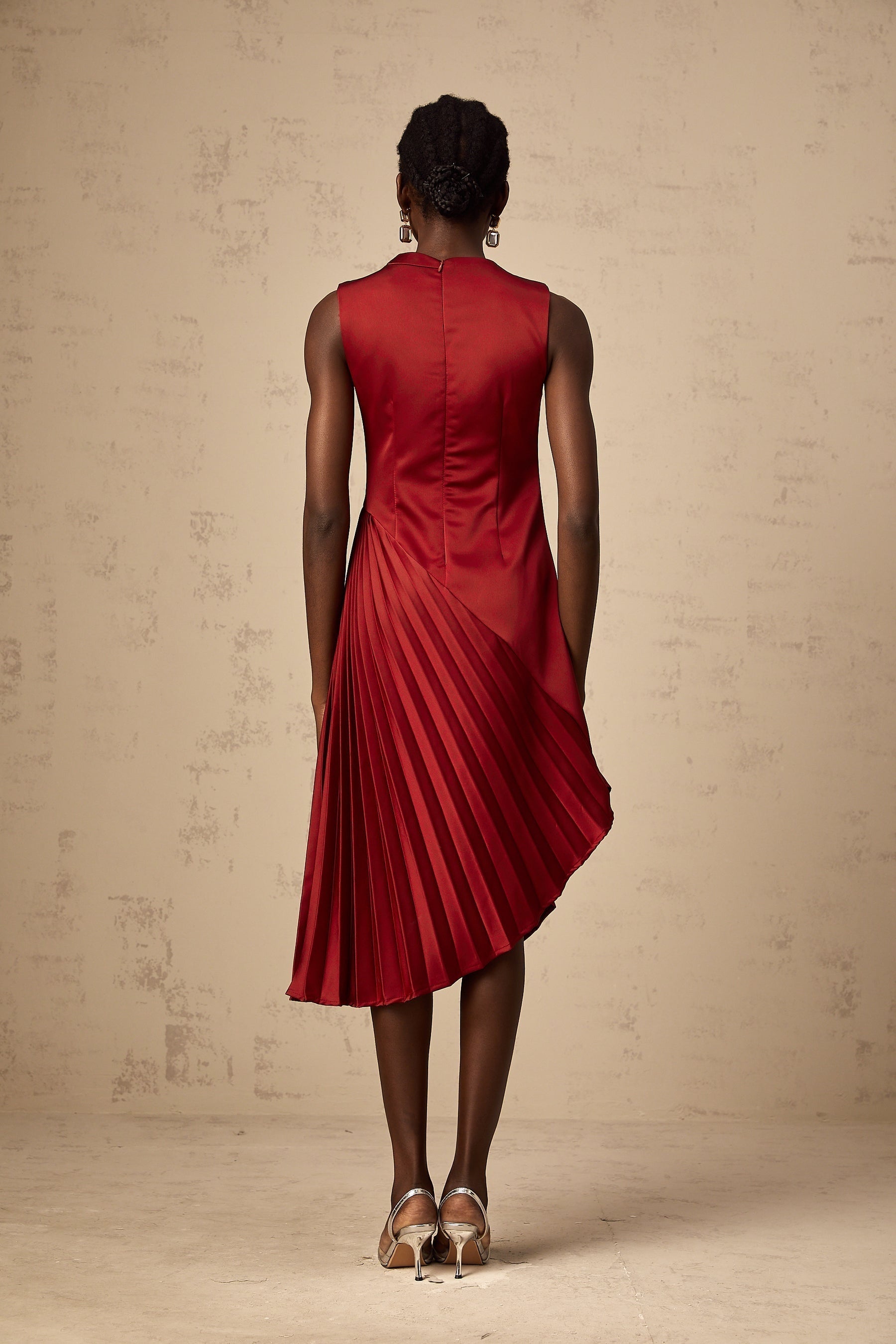 A woman wearing a wine red midi dress with an asymmetric neckline and pleated skirt standing against a plain background