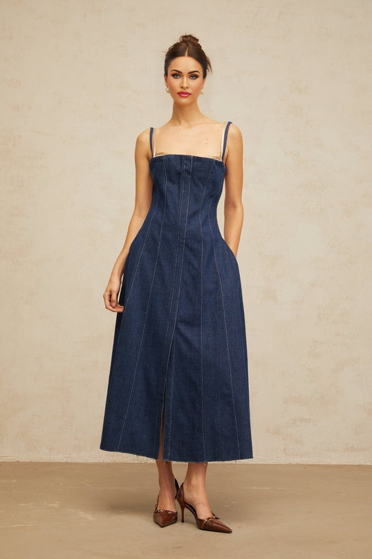 A woman wearing an indigo blue denim midi dress with panel detailing standing against a plain background