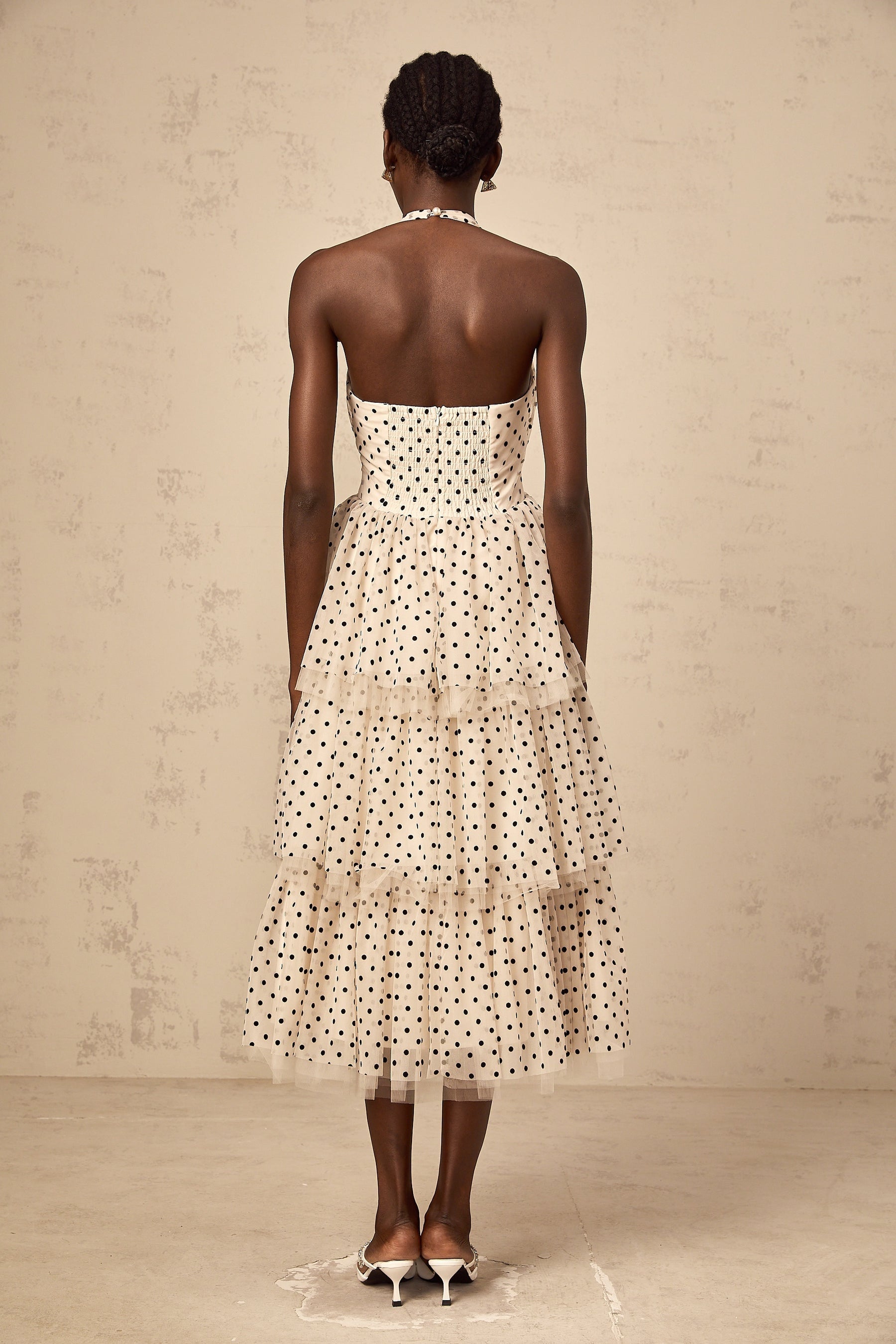 A woman wearing an ivory colored midi dress with polka dots and tiered layers standing in a neutral background