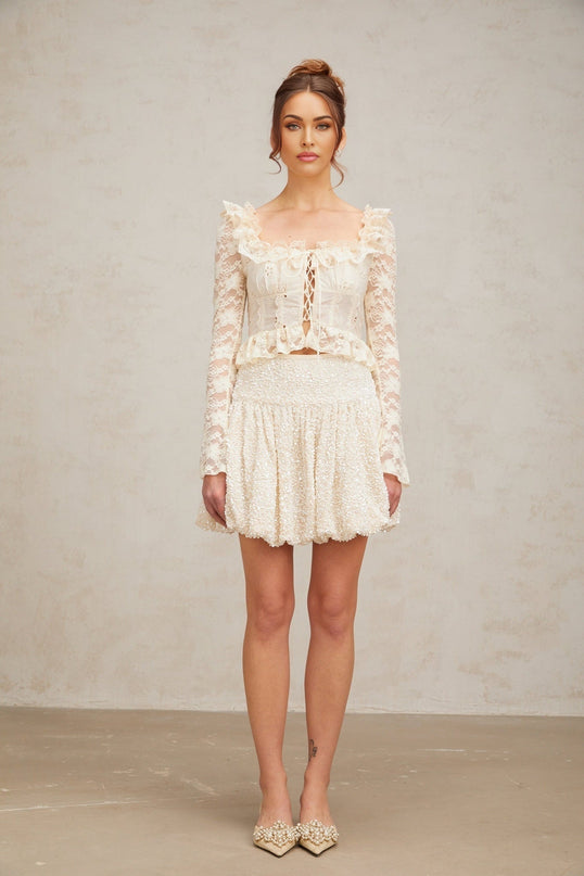 A woman wearing an off white blouse with floral lace detailing and ruffled accents
