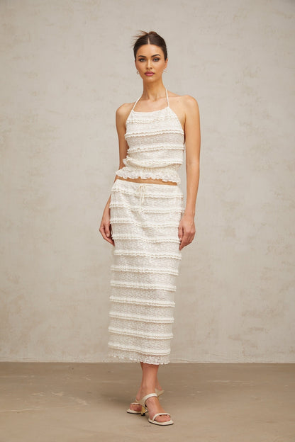 A woman wearing an off white floral lace matching set consisting of a ruffled top and skirt the top has short sleeves and the skirt falls to mid thigh length