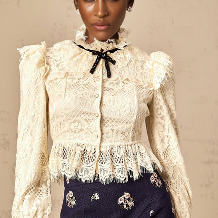 A woman wearing an offwhite blouse with floral lace detailing and a bowknot accent at the neckline