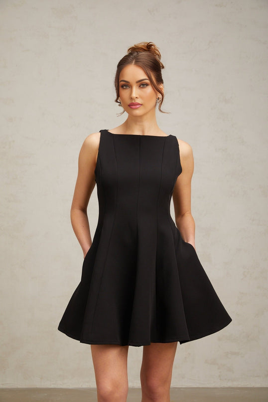 A young woman wearing a black sleeveless mini dress with a flared skirt standing against a plain background