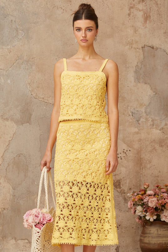 A young woman wearing a bright yellow crochet knit matching set consisting of a sleeveless crop top and a high waisted mini skirt standing against a plain background
