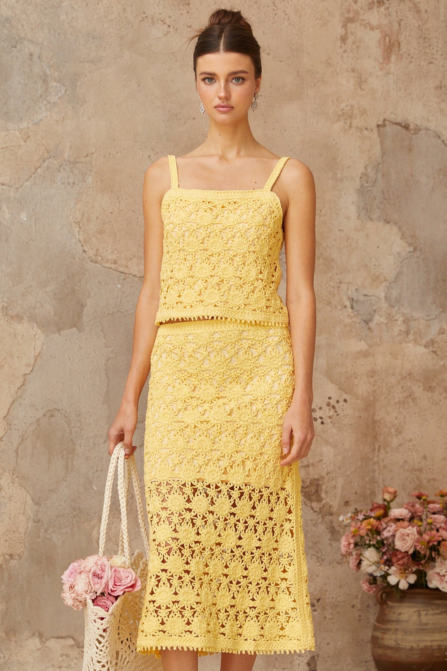 A young woman wearing a bright yellow crochet knit matching set consisting of a sleeveless crop top and a high waisted mini skirt standing against a plain background