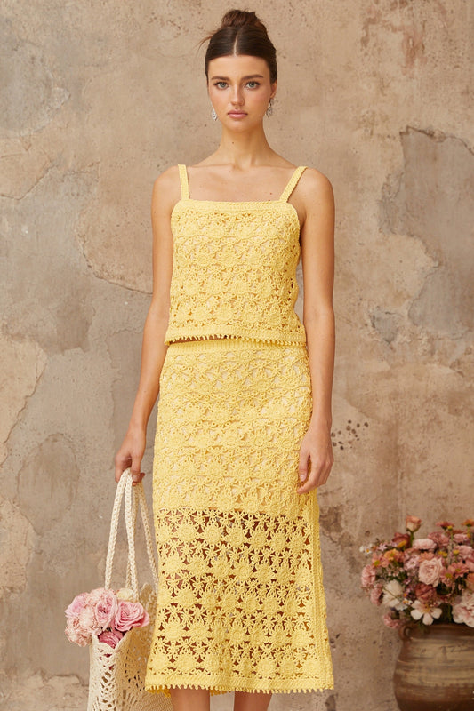 A young woman wearing a bright yellow crochet knit matching set consisting of a sleeveless crop top and a high waisted mini skirt standing against a plain background