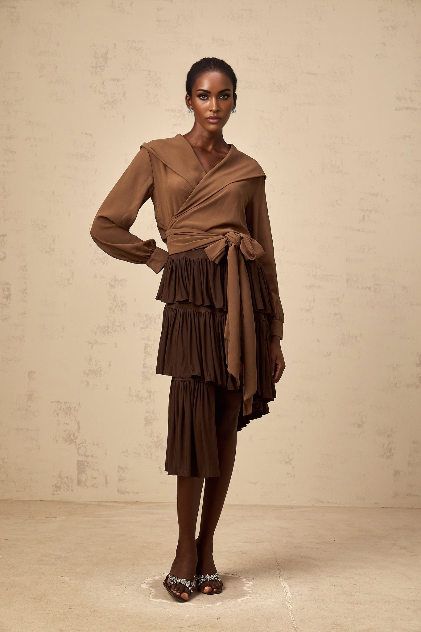 A young woman wearing a brown asymmetrical mini skirt with pleated tiers standing against a plain background