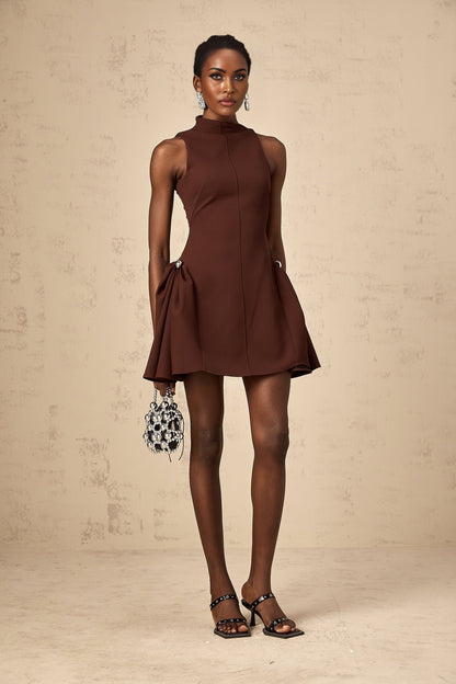 A young woman wearing a brown mini dress with an asymmetric neckline and puffed sleeves standing against a plain background