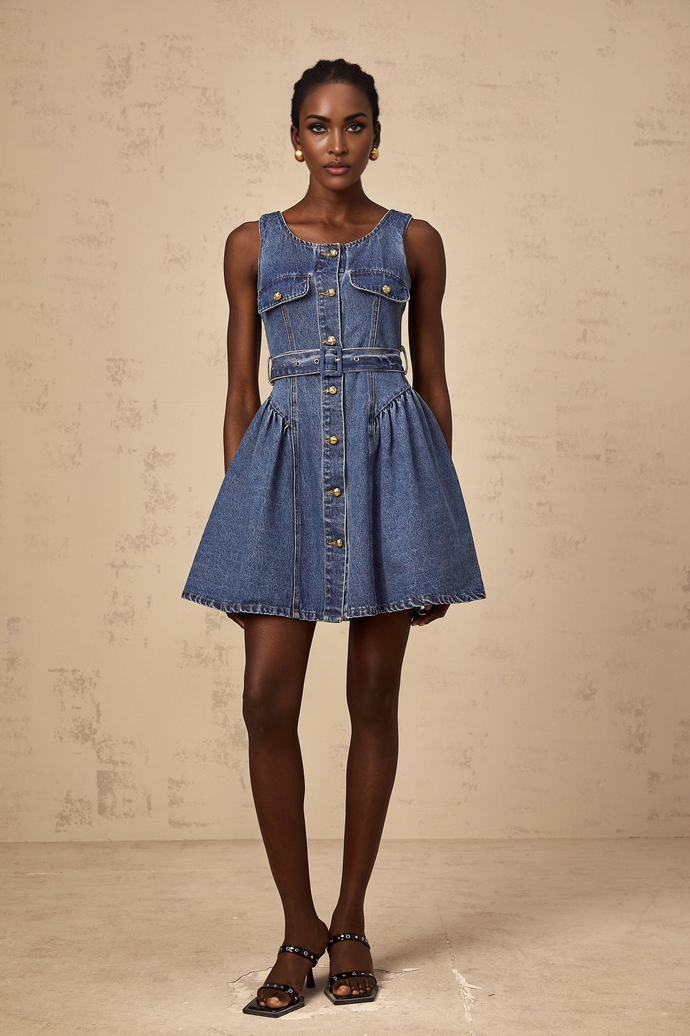 A young woman wearing a light blue denim mini dress with short sleeves and a belted waist standing against a plain background