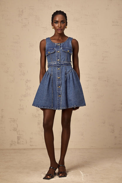 A young woman wearing a light blue denim mini dress with short sleeves and a belted waist standing against a plain background