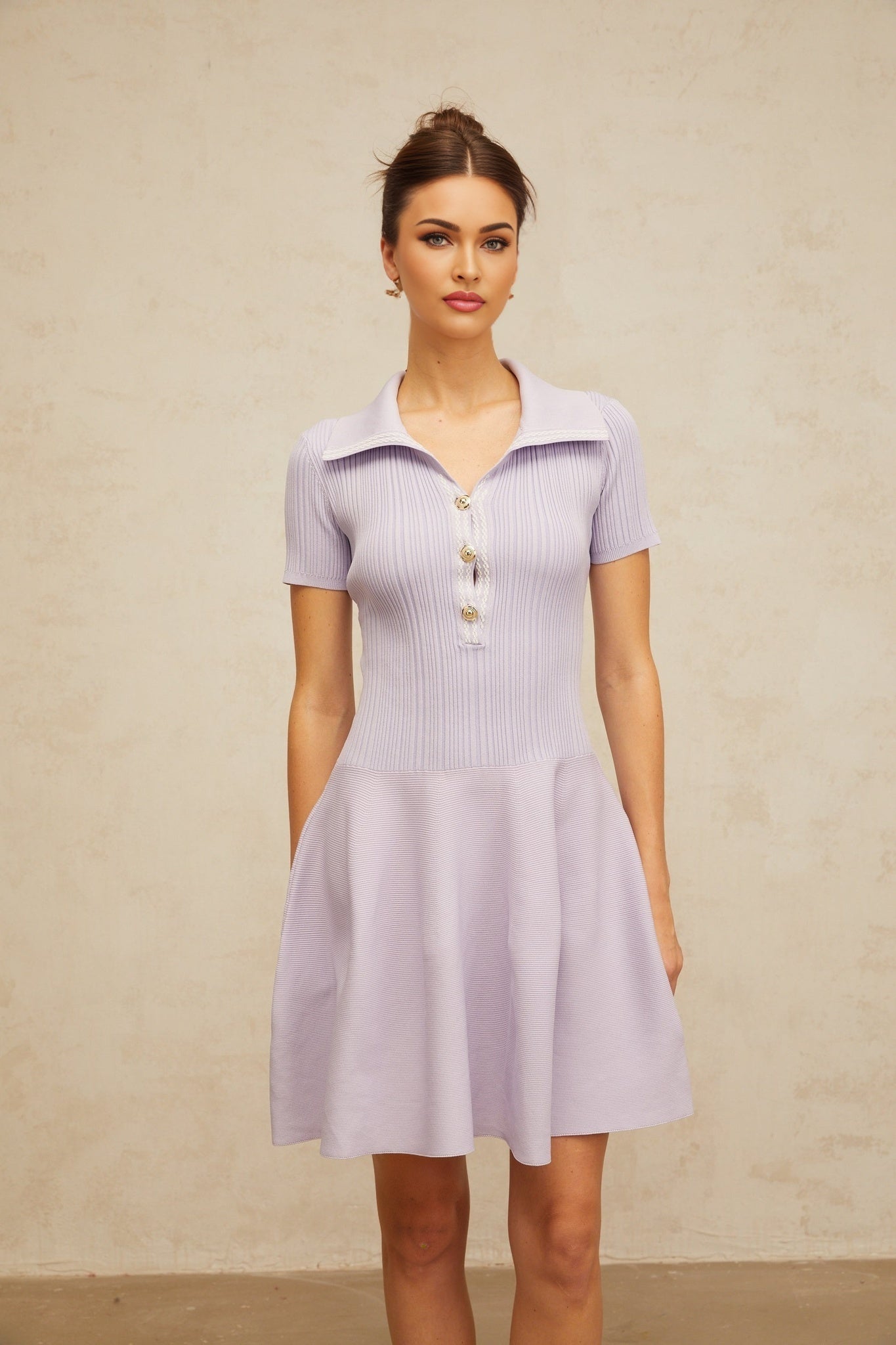 A young woman wearing a light purple knit mini dress with a fitted shirred bodice and flared skirt standing against a plain background
