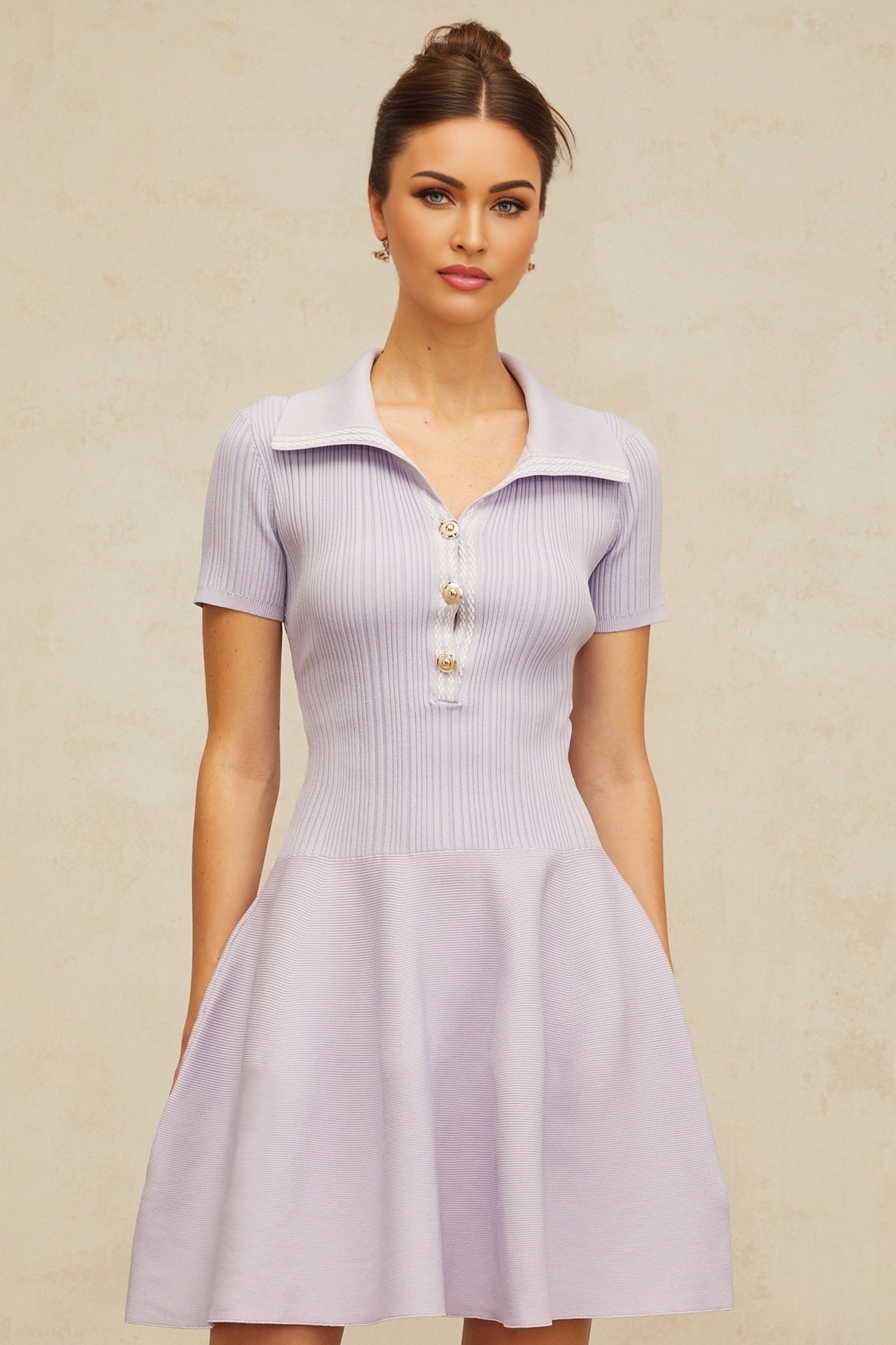 A young woman wearing a light purple mini dress with a fitted bodice and flared skirt The dress features shirred detailing and is made of knit fabric The model stands in a relaxed pose showcasing the dresses flowy silhouette