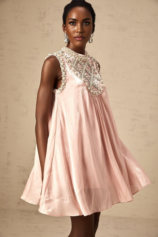 A young woman wearing a short pink dress with pleated skirt and embellished bodice standing against a plain background