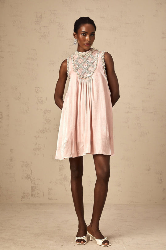 A young woman wearing a short pink mini dress with embellishments and pleats standing in a neutral setting