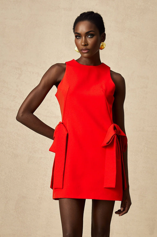 A young woman wearing a short red sleeveless mini dress with a bow detail at the front standing against a plain background