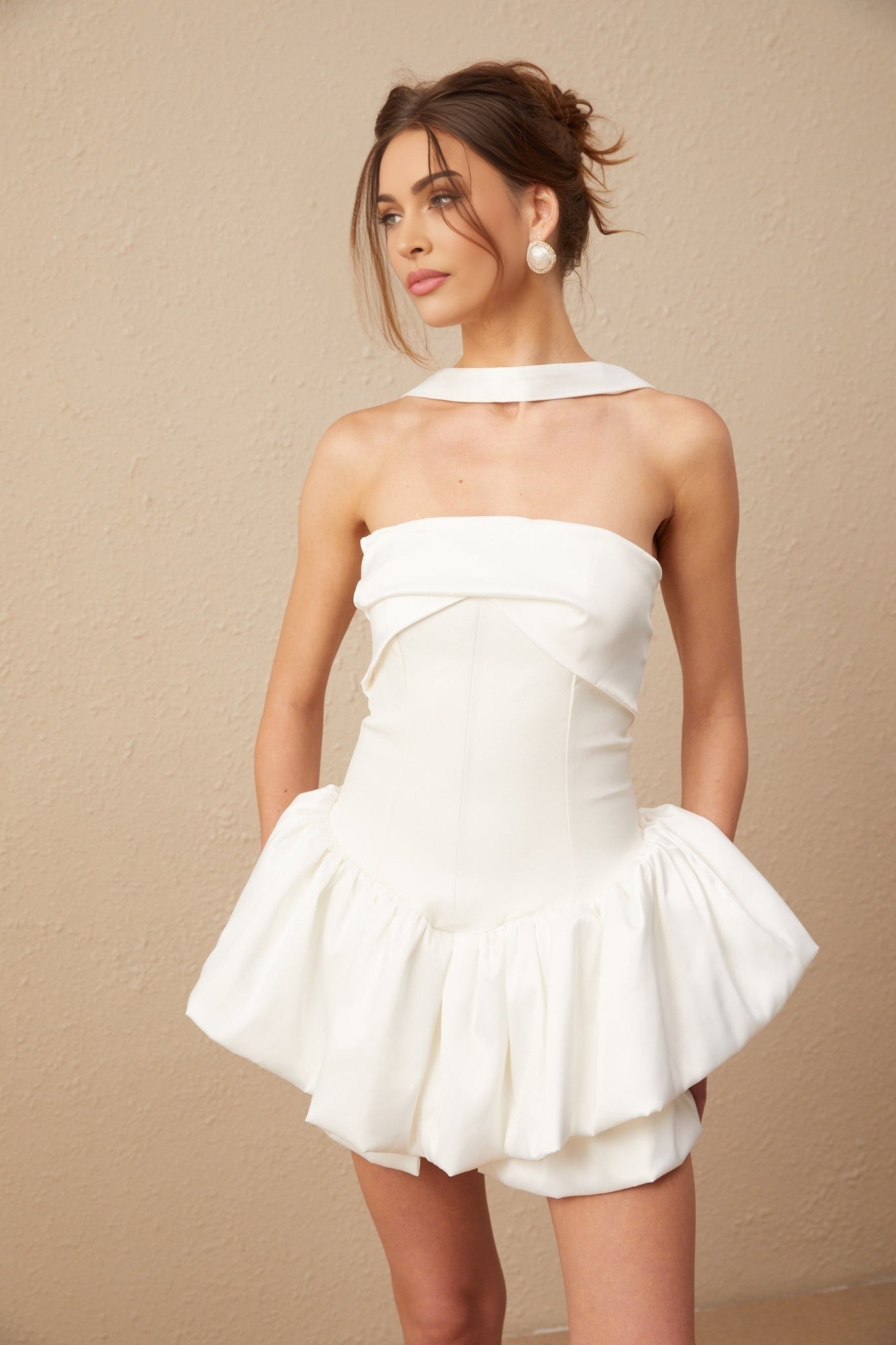 A young woman wearing a short white halterneck dress with ruffled details posing against a plain background