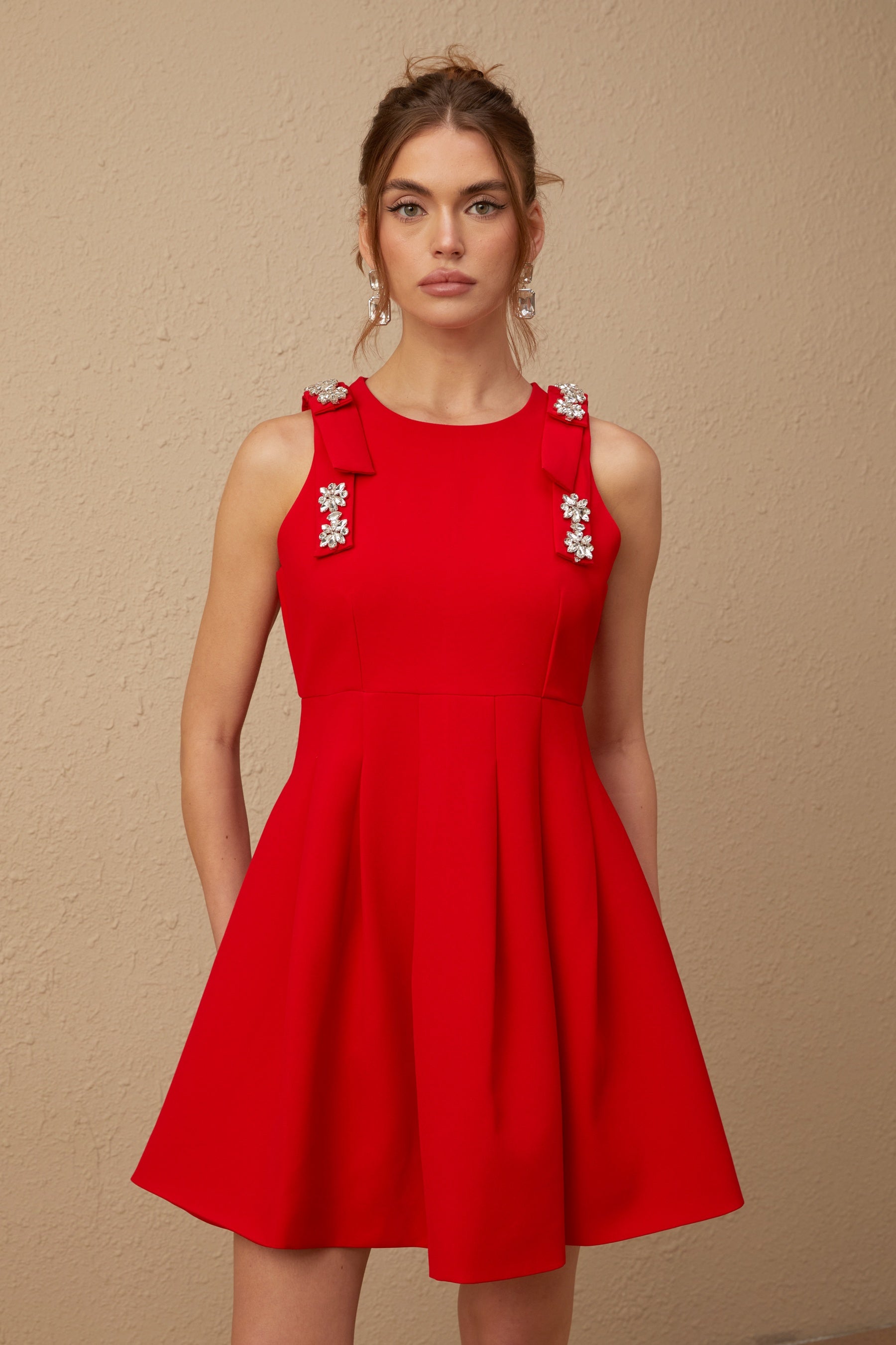 A young woman wearing a sleeveless red mini dress with a pleated skirt and a large bow detail at the neckline
