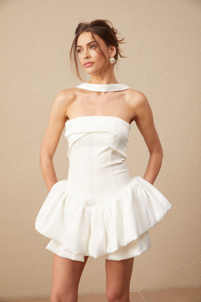 A young woman wearing a white halterneck mini dress with ruffled details standing against a plain background