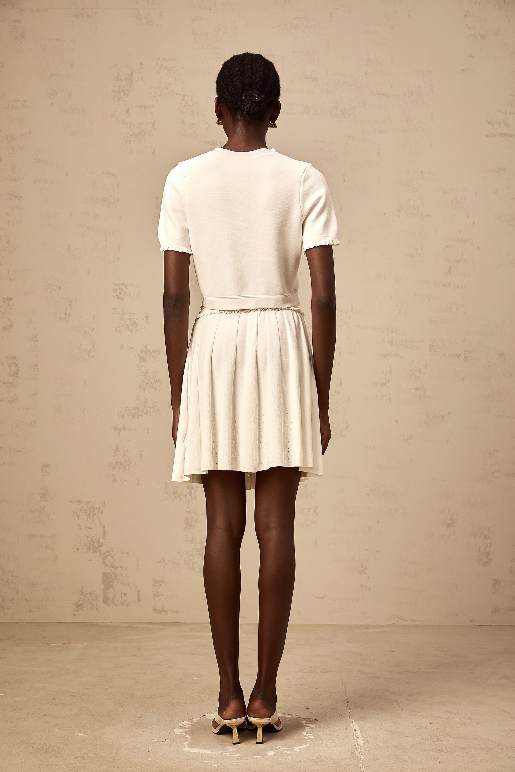 A young woman wearing a white knit top with ruffled trim and a matching white skirt standing in a relaxed pose against a plain background