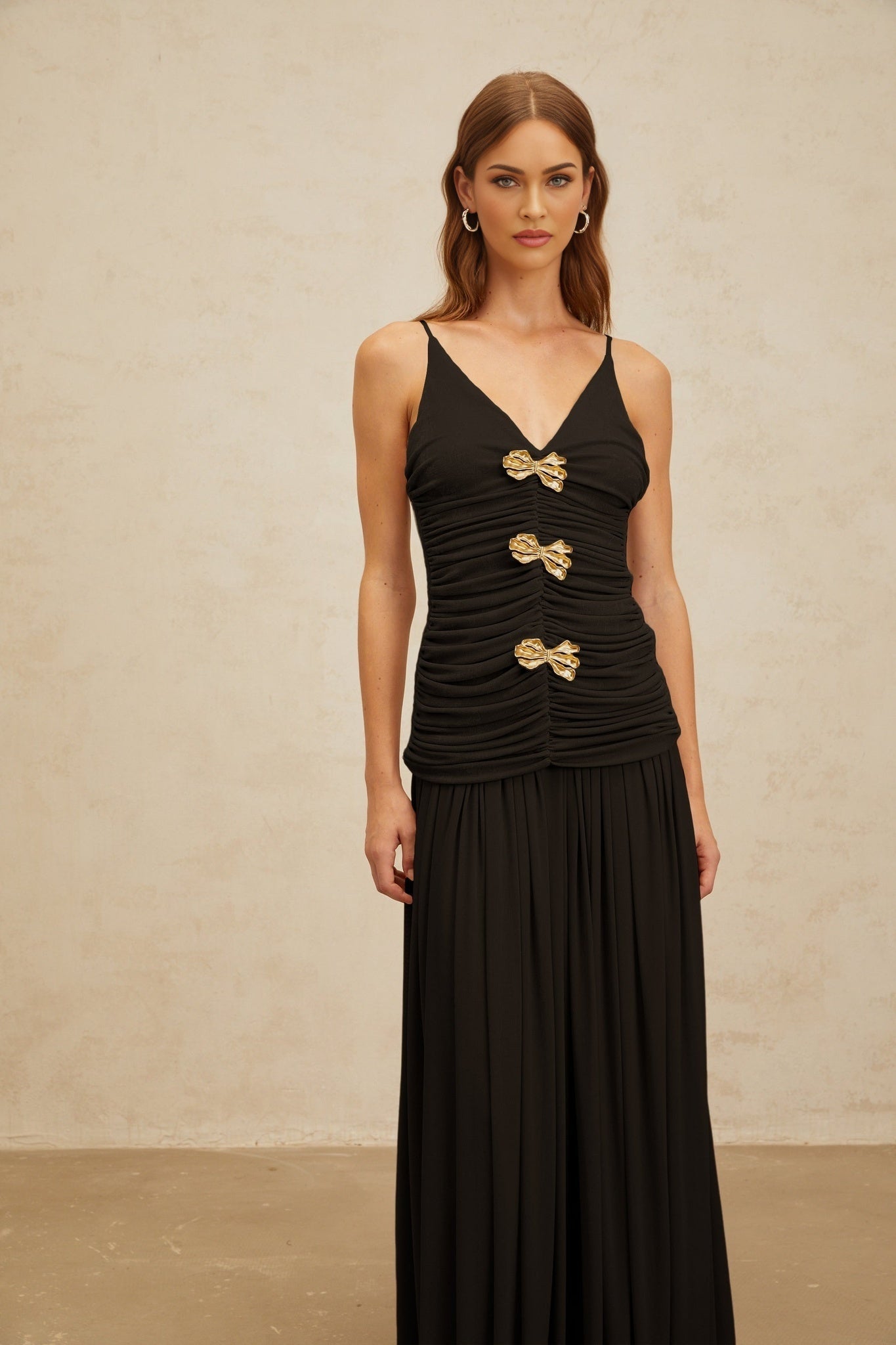 Black V-neck sleeveless top with adjustable spaghetti straps, brooch detailing, and ruched detailing, paired with a black pleated maxi skirt