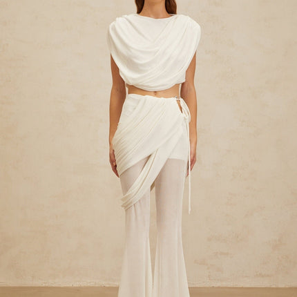 White sleeveless bateau neck top with ruched detailing and cropped length, paired with white high rise trousers with asymmetrical design and flared hem