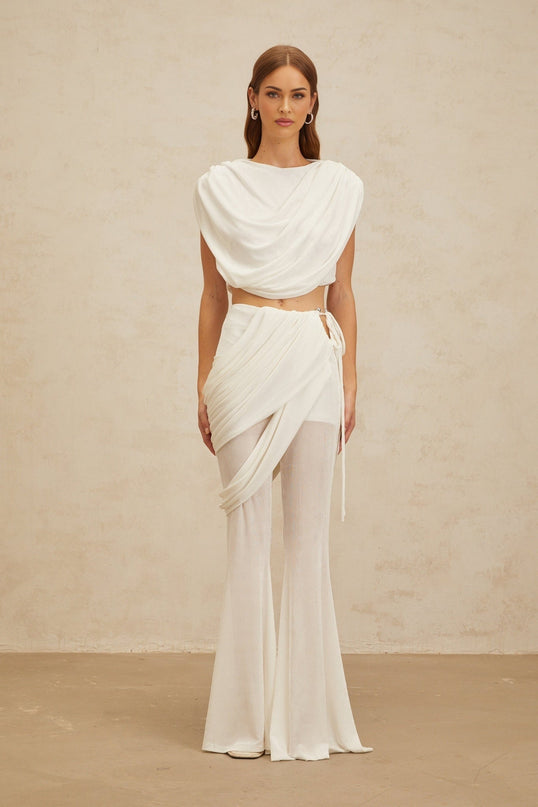 White sleeveless bateau neck top with ruched detailing and cropped length, paired with white high rise trousers with asymmetrical design and flared hem
