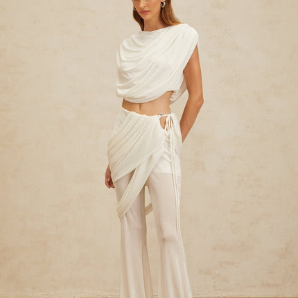 White sleeveless bateau neck top with ruched detailing and cropped length, paired with white high rise trousers with asymmetrical design and flared hem