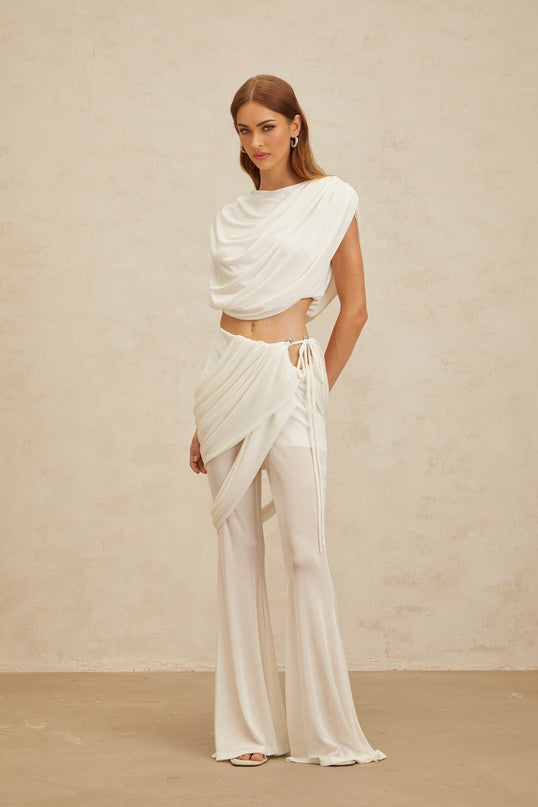 White sleeveless bateau neck top with ruched detailing and cropped length, paired with white high rise trousers with asymmetrical design and flared hem