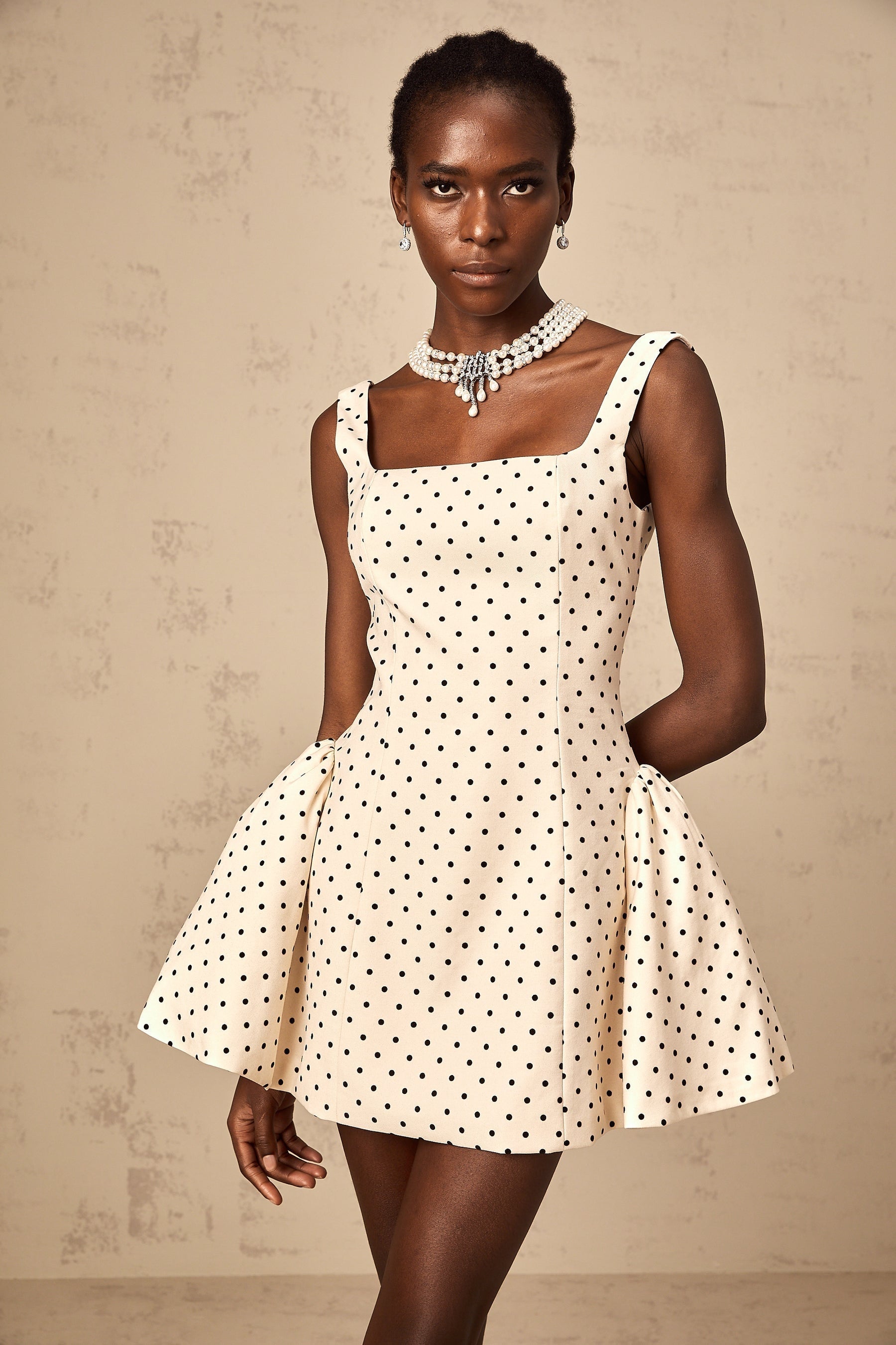 Woman wearing a beige mini dress with white polka dots and puff sleeves standing against a plain background
