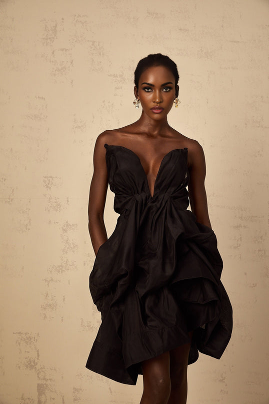 Woman wearing a black asymmetrical pleated cocktail mini dress with one shoulder strap standing against a plain background