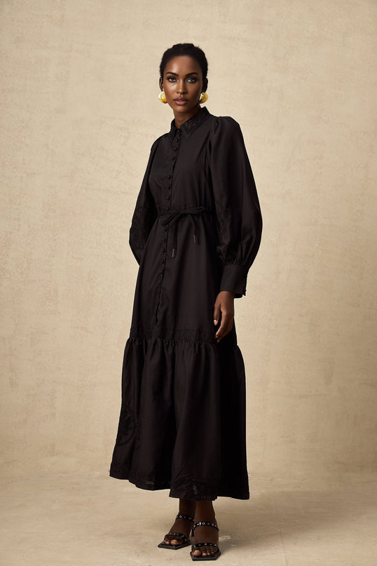 Woman wearing a black lace midi dress with embroidered details and sheer sleeves standing against a plain background