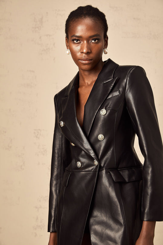 Woman wearing a black leather blazer mini dress with a fitted silhouette and lapel collar standing against a plain background